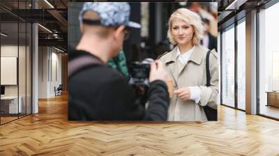 A girl makes a video blog with a cameraman on the street. Young woman blogger, journalist filming with a professional camera and videographer. Wall mural