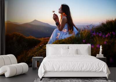 A girl sits on the edge of the cliff and looking at the sun valley and mountains. Woman sitting on mountain top and contemplating the sunset. Young woman in long white dress Wall mural