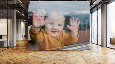 A little happy boy is looking outside through the window from inside the room. View from the street. Sweet toddler child put his hands on the window. Closeup smile on son's face. Funny moments. Wall mural