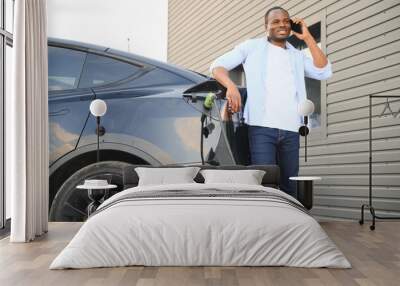 An African man is standing near an electric car, waiting for it to charge at a charging station and using the phone. Wall mural