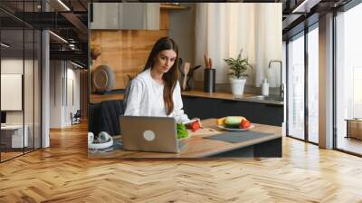 Athletic woman blogger nutritionist prepare a salad with fresh vegetables and conducts a video conference on healthy eating on laptop in the kitchen Wall mural