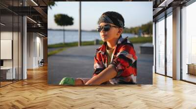 cute boy on roller skates sits on the ground, rests while learning to skate in the park on a summer day Wall mural