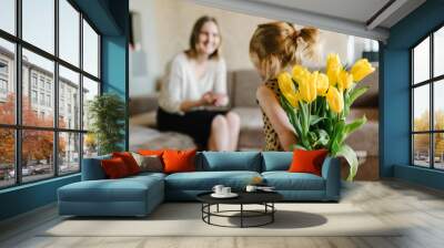 Cute little girl greeting mother and gives her a bouquet of flowers tulips at home. Mother's day concept. Mom and daughter smiling. Happy family holiday and togetherness. Back view. Wall mural