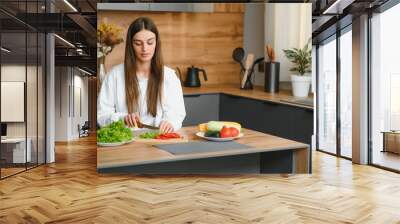 Happy smiling cute woman is preparing a fresh healthy vegan salad with many vegetables in the kitchen at home and trying a new recipe Wall mural