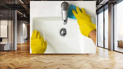 Hygiene concept aerial close up view of female hands wearing yellow protective rubber gloves cleaning a white modern ceramic sink around chrome tap or faucet and fittings using a blue sponge cloth Wall mural