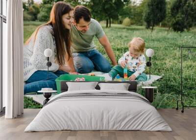 Mom, dad, child 2 year old play constructor game sitting in green grass on blanket sunny summer day. Family with little cute toddler boy playing car toys in garden. Mother, father, son walking in park Wall mural