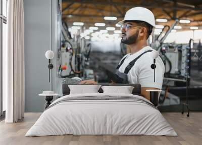 Operator of machine. Industrial worker indoors in factory. Young technician in white hard hat Wall mural