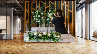 Organizer women arranges and decoration wedding. Setup. Table newlyweds in banquet hall on party. Festive table with arch, decorated with composition of flowers and greenery, candles. Back view. Wall mural