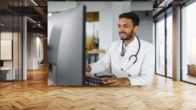 People, occupation and medicine concept. Smiling male Indian Arab doctor in white coat, sitting in medical office at desk with laptop Wall mural