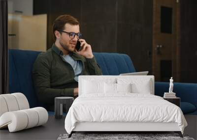 Portrait of an attractive serious young bearded man wearing casual clothes sitting on a couch at the living room, talking on mobile phone while using laptop computer. Wall mural