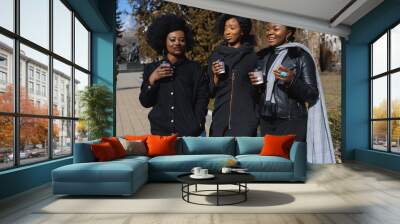 three beautiful and stylish dark-skinned girls with long hair standing in a city and drinking a coffee and use the phones Wall mural