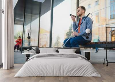 Young blind man with smartphone sitting on bench in park in city, calling. Wall mural