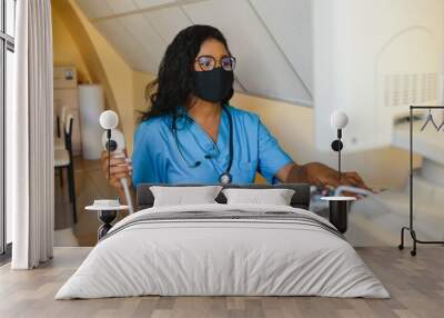 Young cheerful African woman operator in mask of an ultrasound scanning machine analyzing diagnostics results of patient. Young smiling African doctor working on a modern ultrasound equipment. Wall mural