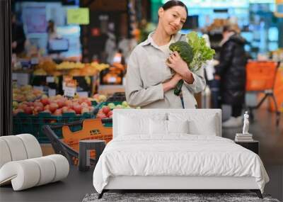 Young woman chooses broccoli, buying vegetables in supermarket. Wall mural