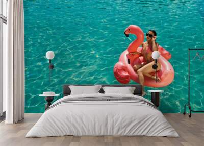 Above view of brunette girl swimming in pool, sitting on pink flamingo. Slim young female in swimsuit and sunglasses relaxing, sunbathing, enjoying. Concept of summertime. Wall mural