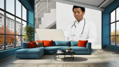 African doctor in coat using laptop on reception. Wall mural