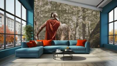 Back view of man in gladiator helmet and red long cloak standing with round shield and spear in hands looking away in woods. Spartan. Wall mural