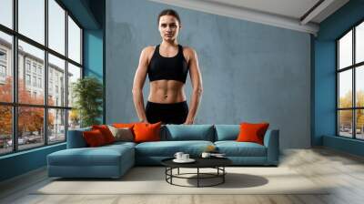 Front view of muscular pretty woman wearing black sports underwear and carrying dumbbells. Isolated portrait of pumped fitnesswoman posing on gray studio background, loft interior. Concept of sport. Wall mural