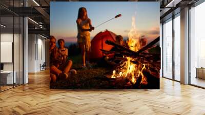 Happy teen girl frying sausage over fire, having fun at campsite at dusk. Side view of group of kids sitting around big campfire, roasting meal at twilight against colorful sky. Concept of camping. Wall mural