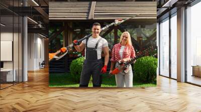 Portrait of happy caucasian man and woman posing at beautiful garden with electric hedge trimmers in hands. Male and female gardeners enjoying seasonal work with plants. Wall mural