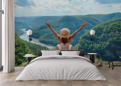 A female traveler sits with open arms on a mountaintop, overlooking a winding river and lush green forested landscape Wall mural