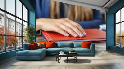 A hand with neatly trimmed nails rests on a red book adorned with Cyrillic text, suggesting formality and importance. The wooden surface and blurred background create a calm, contemplative atmosphere, Wall mural