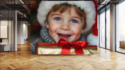 A joyful young child wearing a Santa hat smiles brightly while holding a beautifully wrapped Christmas present. The warm indoor atmosphere is enhanced by a decorated tree in the background, evoking ho Wall mural