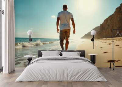 A man in shorts and a white T-shirt is seen walking barefoot along a sandy beach during a sunny day, leaving a trail of footprints behind him, with waves lapping at the shore and rock formations nearb Wall mural