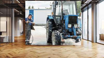 A man is washing a tractor with a pressure washer. The tractor is blue and has a large tire Wall mural