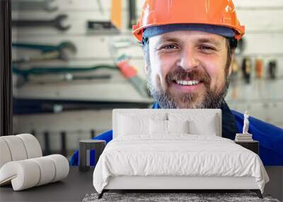 A man with a beard in a helmet and work clothes is smiling a snow-white smile against the background of a stand with tools. Portrait of a worker in workwear with copy space. Wall mural