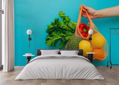 A hand holding an orange net bag with vegetables and fruits on a blue background. Wall mural