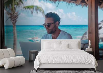 A young man in a T-shirt works on a laptop at a table on the beach in the summer. Wall mural