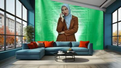 angry young Asian woman wearing hijab and blazer receiving incoming call on smartphone on green background
 Wall mural