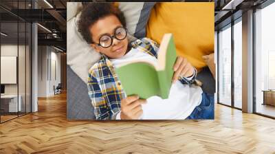 Above view portrait of cute African boy wearing big glasses reading book while lying on couch, homeschooling concept, copy space Wall mural