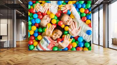 Above view portrait of three happy little kids in ball pit smiling at camera raising hands while having fun in children play center, copy space Wall mural