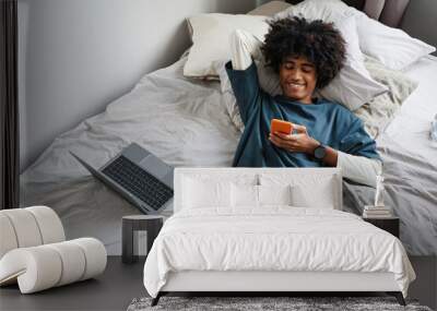 Above view portrait of young African-American man lying on bed at home and smiling while using smartphone, copy space Wall mural