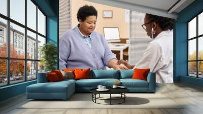 African American senior woman dressed in sweater sitting in clinic office with closed eyes while female doctor taking her pulse rate by hand Wall mural