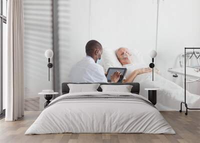 Back view portrait of African-American doctor using digital tablet while sitting by senior patient in hospital bed, copy space Wall mural