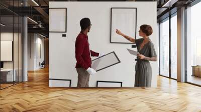 Back view portrait of female art gallery manager instructing worker hanging painting frames on white wall while planning exhibition in museum, copy space Wall mural