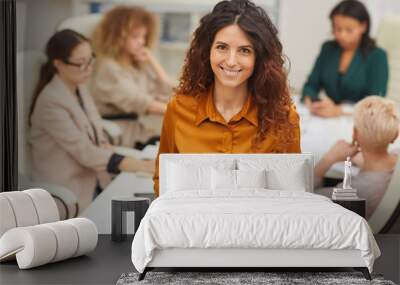 Charming Caucasian woman standing with with eyeglasses taken off looking at camera smiling with colleagues coworking behind her medium portrait Wall mural