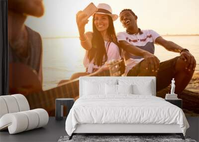 Cheerful beautiful Hispanic girl taking selfie with African boyfriend while they sitting on log and listening to street musician on beach Wall mural