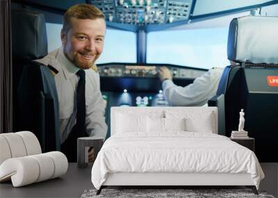 Cheerful confident bearded pilot in white shirt and black tie sitting in armchair and turning back to look at camera in cockpit Wall mural