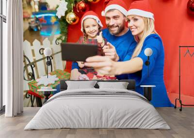 Cheerful modern family with daughter sitting on bench in mall with Christmas gifts making selfie Wall mural