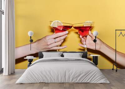 Close up of two female hands holding red cocktails against vibrant yellow background, copy space Wall mural