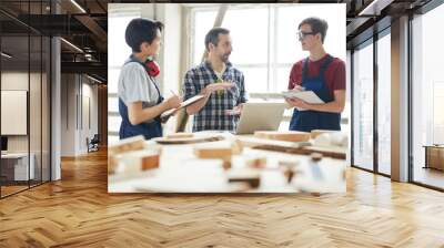 Confident handsome chief carpenter in checkered shirt gesturing hands while giving class for interns in workshop, young students making notes in sketchpads Wall mural