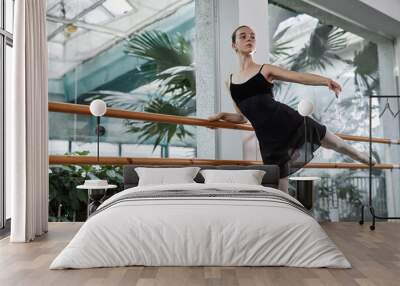 Confident teenage ballet dancer gracefully performing Arabesque position standing on one leg at barre in dance studio with huge windows, copy space Wall mural