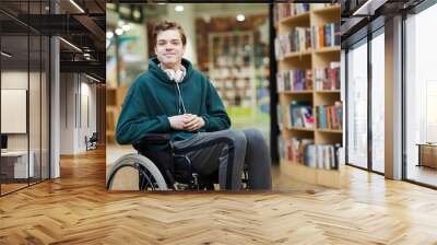 Content handsome young disabled student with headphones on neck siting in wheelchair and looking at camera in modern library or bookstore Wall mural