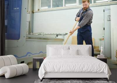 Content handsome young man in workwear using broom while sweeping floor in dusty factory shop Wall mural