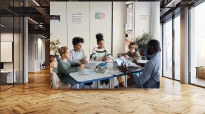 diverse group of children sitting at table with male teacher in modern school classroom Wall mural