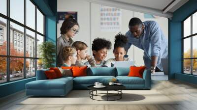 Diverse group of children with male teacher using laptop together in modern school classroom Wall mural